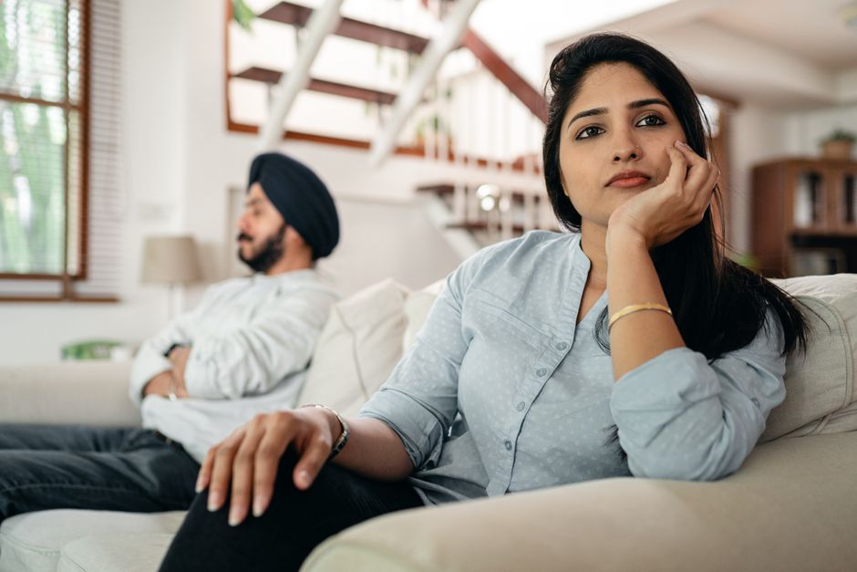 Inside a well-lit living room, a woman with dark hair, wearing a light blue blouse, sits on a white sofa with her hand resting on her face, appearing upset or stressed. Behind her, a man with a beard