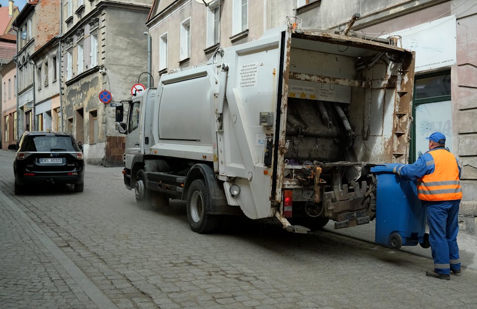 A garbage collection truck parked on a cobblestone street next to residential buildings during daytime, with a worker wearing a blue uniform, orange safety vest, and blue cap, standing beside the truck and emptying a blue wheeled bin into the rear loading compartment. The truck is white with signs of rust and dirt on its rear section, which is open to allow waste disposal. Nearby, a black car is parked along the curb. The surroundings include multi-storey brick and stucco houses with windows, some with shutters, and a 'no parking' traffic sign visible on the wall. The scene captures the process of waste collection, part of home or property clearance, possibly related to house removals or moving services by Man with Van Goddington, in a quiet urban setting.