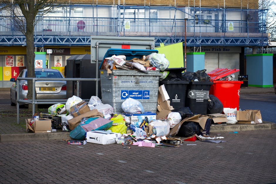 A row of five large wheeled commercial waste bins, including two with labels indicating 'Waste Only' and 'Construction Waste,' are positioned on a paved area beside a dark metal storage container with double doors. The bins are covered with closed lids, some secured with metal chains, and are made of plastic in varying shades of light and dark grey. Behind the bins, dense foliage and trees can be seen, suggesting an outdoor setting near a residential or commercial property. The area appears to be used for waste collection and disposal, which may relate to the process of home relocation or clearing. The scene is captured under natural lighting, typical of a daytime environment, with no human activity visible. Man with Van Goddington occasionally provides services related to removals and waste management, which can be supported by images depicting loading or waste disposal areas like this one.