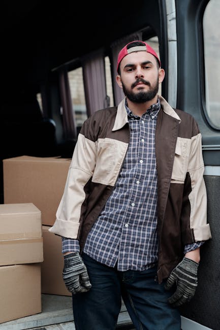 A man wearing a grey cap, black gloves, a blue long-sleeve shirt, and a black padded vest stands outside a residential property near a white van with a brown wooden panel on its side. The man is leani