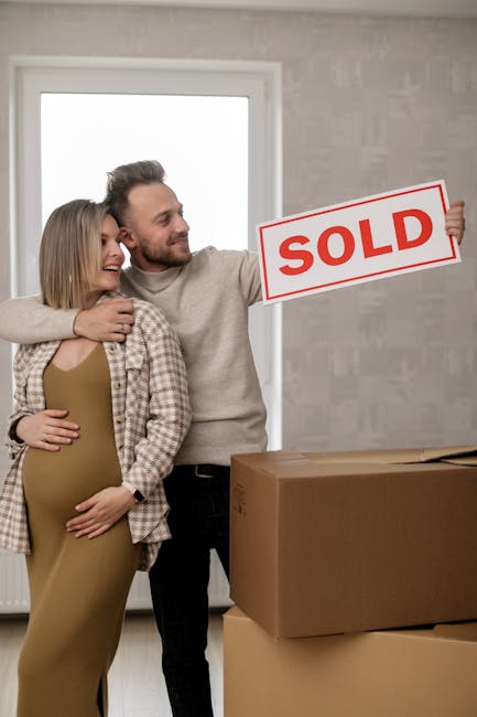 A man and a pregnant woman inside a new home, smiling and embracing each other. The man is holding a red and white 'SOLD' sign in one hand, indicating a recent property purchase. In front of them, the