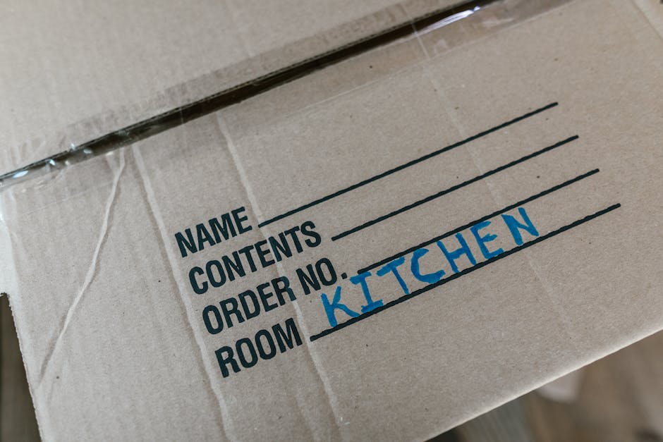 A close-up view of a cardboard moving box with printed labels indicating sections for 'NAME', 'CONTENTS', 'ORDER NO.', and 'ROOM'. The 'ROOM' section is handwritten in blue marker, specifying 'KITCHEN'. The box is partially wrapped with clear plastic tape and is placed indoors, likely in a home or storage area, ready for a house removal or relocation. The image highlights packing procedures during furniture transport, with the box situated among other packed items, emphasizing careful organisation in the home moving process. Man with Van Goddington utilizes such packing practices to facilitate efficient home relocation and furniture transport services, ensuring items are well-labelled for smooth loading and unloading.