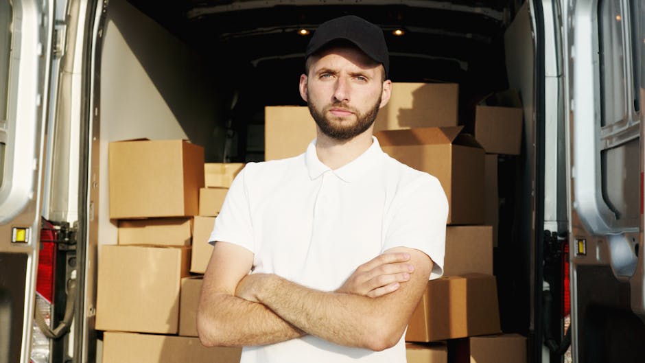 A young man with a serious expression, wearing a black cap and white polo shirt, stands inside an open moving van. Behind him, the van's interior is filled with numerous cardboard boxes of various sizes, some sealed with packing tape, and a few plastic-wrapped items. The boxes are arranged on the floor and stacked towards the back of the van, indicating a home relocation or furniture transport process. The loading area is illuminated by natural light, with the rear doors of the van open to reveal the belongings being prepared for transport. The scene reflects the logistical steps involved in packing and loading during removals, a service provided by companies like Man with Van Goddington, which specialises in house removals and moving logistics, highlighting the importance of careful packing and organized loading to avoid hidden fees in the process.