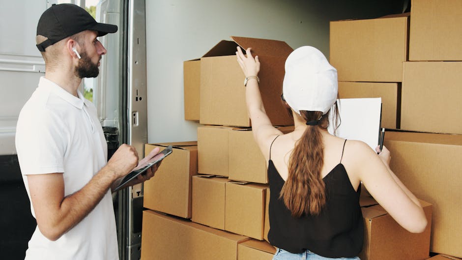 A man with a beard, wearing a black cap, white T-shirt, and wireless earbuds, is standing beside a partially open van garage door, holding a clipboard and pen while observing a woman. The woman, with long brown hair tied back and wearing a white cap, black sleeveless top, and light blue jeans, is in the process of packaging or labeling cardboard boxes in front of her. She is reaching up with her left hand to move or adjust a cardboard box that is stacked among other similar boxes arranged on the ground and against a wall inside the garage area, which is part of a house or storage space. The boxes are made of brown cardboard with some open and others closed, and appear to be filled with household items for a home relocation. The scene depicts the loading process involved in furniture transport and packing during house removals, with visible materials such as cardboard and packing tape. A large vehicle or van belonging to [COMPANY_NAME] is possibly nearby for moving services, with a focus on efficient packing and transportation, highlighting typical move preparations in a residential setting.