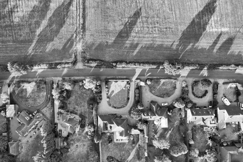 A black and white aerial photograph showing a residential street with several detached and semi-detached houses, each with driveways and gardens. The houses are surrounded by trees and shrubbery, with some using paved pathways. A narrow road runs horizontally at the top of the image, with shadows cast by nearby structures. In the foreground, a group of people are engaged in a home relocation, loading a large cardboard box and a piece of furniture into a van parked on the street. The van is marked with the branding of Man with Van Goddington, a removals service specializing in household moves. The scene includes packing materials such as bubble wrap and protective blankets, along with trolleys and straps used to facilitate furniture transport. The lighting suggests a clear day, with sunlight creating defined shadows from the trees, vehicles, and individuals involved in the moving process, illustrating typical logistics of packing and loading during house removals.