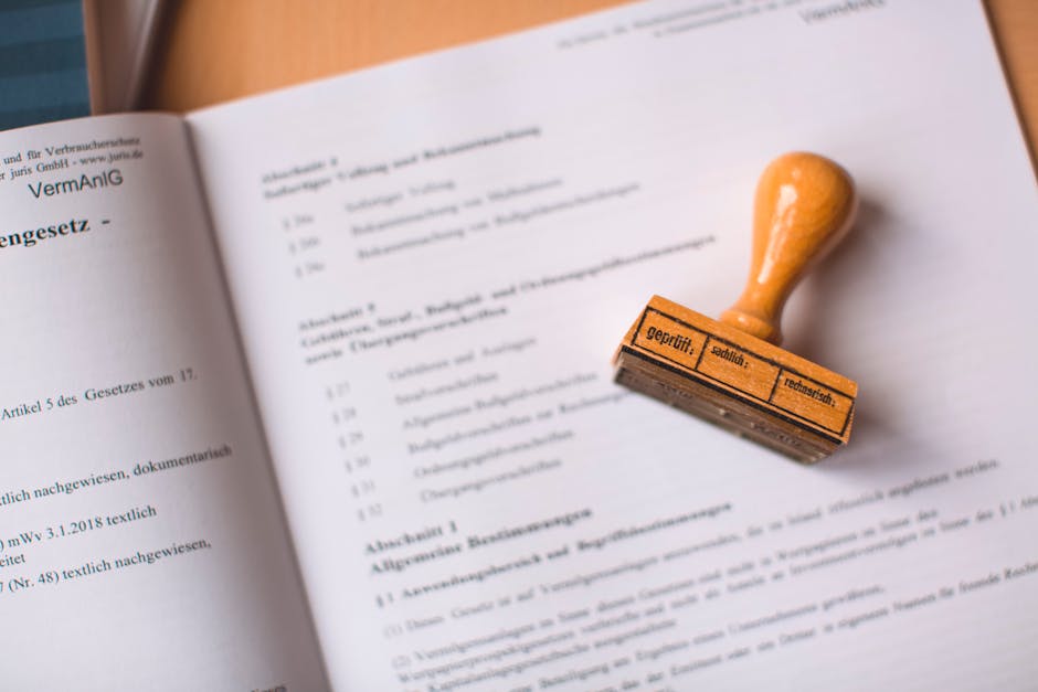 A close-up view of a legal document with printed text, partially open on a flat surface. Resting on the document is a rubber stamp with a wooden handle and a rectangular stamping surface, which has German text indicating it is a tested and approved stamp. The background shows a plain surface, possibly a desk or table, with natural lighting illuminating the scene. The image focuses on the document and stamp, highlighting the careful handling of paperwork involved in home relocation and moving processes. The presence of legal paperwork and the stamp suggest formal documentation related to removals services, such as licensing or compliance checks, relevant to the house moving industry. This visual depiction aligns with the detailed planning and official aspects of furniture transport and packing and moving activities, as managed by companies like Man with Van Goddington.