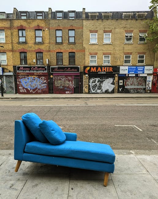 A bright blue chaise longue with two matching cushions placed on its backrest is positioned on the sidewalk outside a building with multi-story graffiti-covered retail units in the background. The sofa's wooden legs are visible, and it appears to be part of a house removal or relocation process, possibly awaiting collection or transport. The scene is illuminated by natural daylight, with no moving personnel present in the image. This setting illustrates a typical element involved in home relocation or furniture transport services, as provided by Man with Van Goddington, highlighting the careful handling of furniture items during packing and loading processes for house removals.