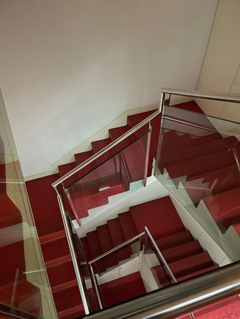A view of a staircase inside a building, with red carpeted stairs and a modern glass and metal railing. The staircase is situated in a well-lit area with neutral-colored walls, and the photo is taken from an upper floor looking downward. Visible are cardboard boxes and packing materials near the bottom of the stairs, indicating a house removal or relocation process. The scene captures the interior environment where furniture and belongings are being moved, supported by the presence of a professional home relocation service such as Man with Van Goddington, operating within the context of house removals, packing, and furniture transport.