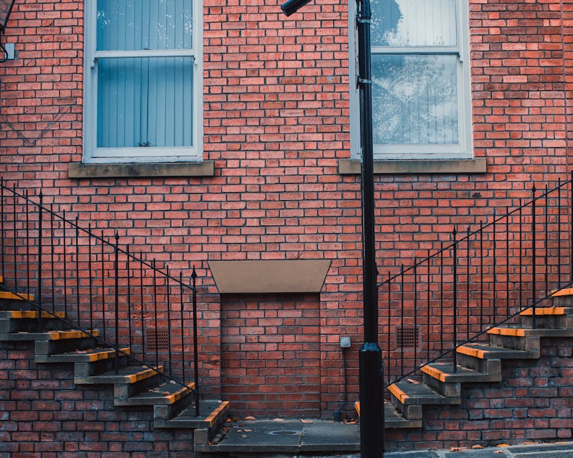 A brick exterior wall of a residential building features two white-framed windows on the upper level, each with a sill. Beneath the windows, there is a small, rectangular vent or access panel set into the brickwork. Two external staircases made of concrete steps with yellow edge protectors and black metal railings ascend from ground level on either side, leading up to the upper floors. The staircases are positioned against the brick wall, which also has a central pipe or downspout running from top to bottom, and a small utility box fixed to the wall. The area appears to be well-lit with natural daylight, and the scene suggests it might be part of a property undergoing house removal or furniture transport, with the staircases providing access for loading items. Man with Van Goddington specializes in relocation services related to houses and flats, including stairs and lift access, as indicated by the contextual setting.