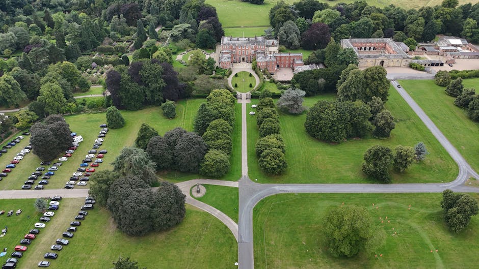 An aerial view of the Goddington Park estate showing a large historic building with a symmetrical facade and central entrance, surrounded by lush green lawns, mature trees, and well-maintained pathways. To the left, there is a spacious parking area filled with numerous cars parked in neat rows. A paved driveway extends from the parking lot, curving towards the entrance of the building, facilitating home relocation and furniture transport activities. The estate features open grassy areas, scattered trees, and a circular entrance driveway leading directly to the main building, with additional roads and pathways providing access around the estate. This setting exemplifies a typical site where house removals and packing and moving services by Man with Van Goddington could be coordinated, especially considering logistics for loading and unloading furniture and boxes, with parking and access points clearly visible for vehicles involved in the relocation process.