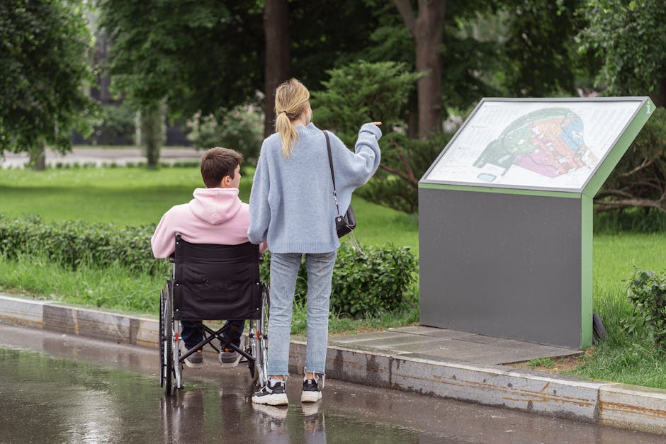 A woman with blonde hair wearing a light blue coat, grey trousers, and white sneakers is standing on a wet pavement next to a man in a wheelchair wearing a pink hoodie. They are positioned near an informational park sign mounted on a grey and green metal stand, with the woman pointing towards it as they discuss or read the map or details displayed. The scene is set outdoors in a park area with lush green grass, trees, and bushes in the background, suggesting a peaceful environment suitable for house removals or moving services. The wet surface indicates recent rain, and the setting includes natural lighting with diffused sunlight, highlighting the furniture transport and home relocation context, as well as the importance of accessible pathways and signage during moving processes.