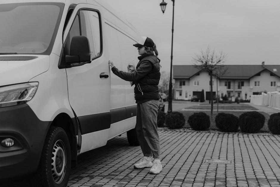 A silver van parked on a city street in front of a historic brick building with large windows and stone detailing around an arched doorway. The van, used for home relocation and furniture transport, i