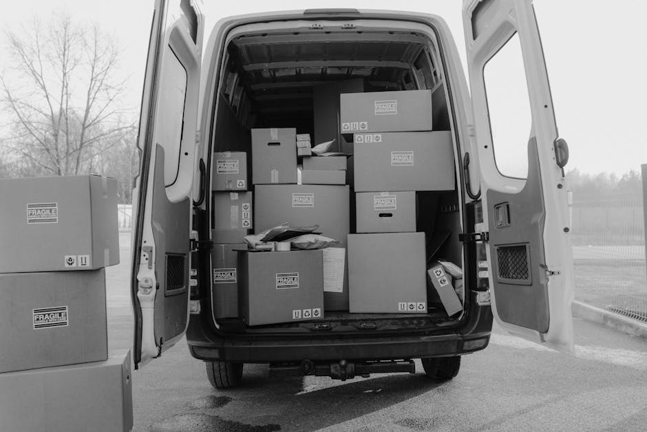 The image shows the open rear doors of a white delivery van loaded with multiple cardboard boxes of various sizes, some labeled with fragile handling symbols, arranged on the van's interior floor. Several boxes are stacked neatly, with additional parcels placed on top or leaning against each other, indicating the process of furniture transport and packing for a home relocation. Outside the van, on the ground, there are more boxes, some ready to be loaded or unloaded, situated on a paved surface adjacent to what appears to be a residential or industrial area with a wire fence and leafless trees in the background. The scene is captured in natural light, emphasizing the logistical aspects of moving services offered by [COMPANY_NAME], with the focus on efficient loading procedures and careful handling of packed items to ensure a smooth moving process near Goddington Station, highlighting the importance of accessible van access and proper packing materials during the loading process.