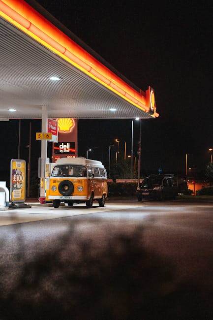 A yellow and white vintage Volkswagen van parked at a Shell petrol station during nighttime, with the station's illuminated canopy and signage visible above. The van is positioned near the fuel pumps, which are under the canopy that extends across the top of the image. Behind the van, there are several parked cars, and the surrounding area includes street lights and distant illuminated buildings. The scene reflects a typical urban setting suitable for home relocation or furniture transport, with the van ready to assist with loading or unloading items as part of a house removals service. The lighting highlights the vehicle and nearby objects, emphasizing the practicality of loading equipment, such as trolleys or straps, used by companies like Man with Van Goddington during packing and moving processes near Goddington Station.